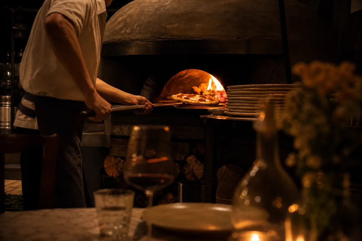 Four à bois de pizzeria vu depuis la salle, pizzaïolo enfournant à la pelle